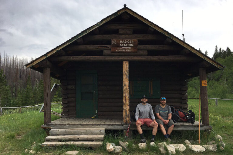 Two hikers sitting on the porch of a log cabin