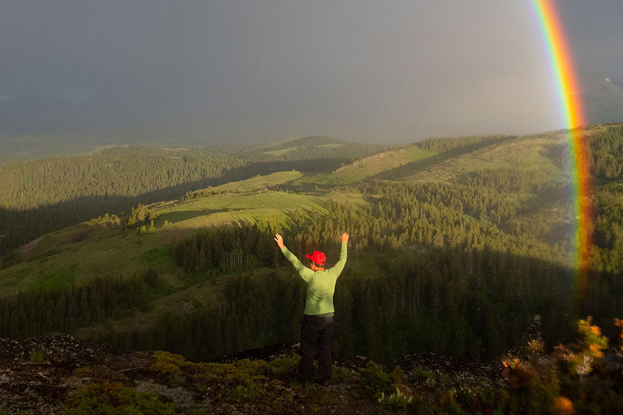 A hiker standing at a vista, arms raised, under a rainbow